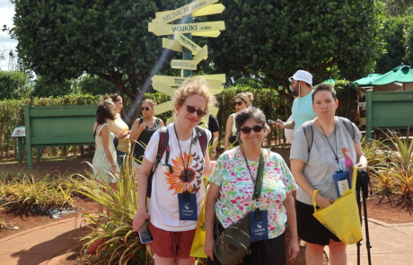 Highlight Vacations travelers standing in front of pineapple plant with a sign post (with arrows point to different locations in the world)