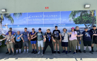 Travelers in front of sign and saying "we're here in front of the Cayman Islands sign